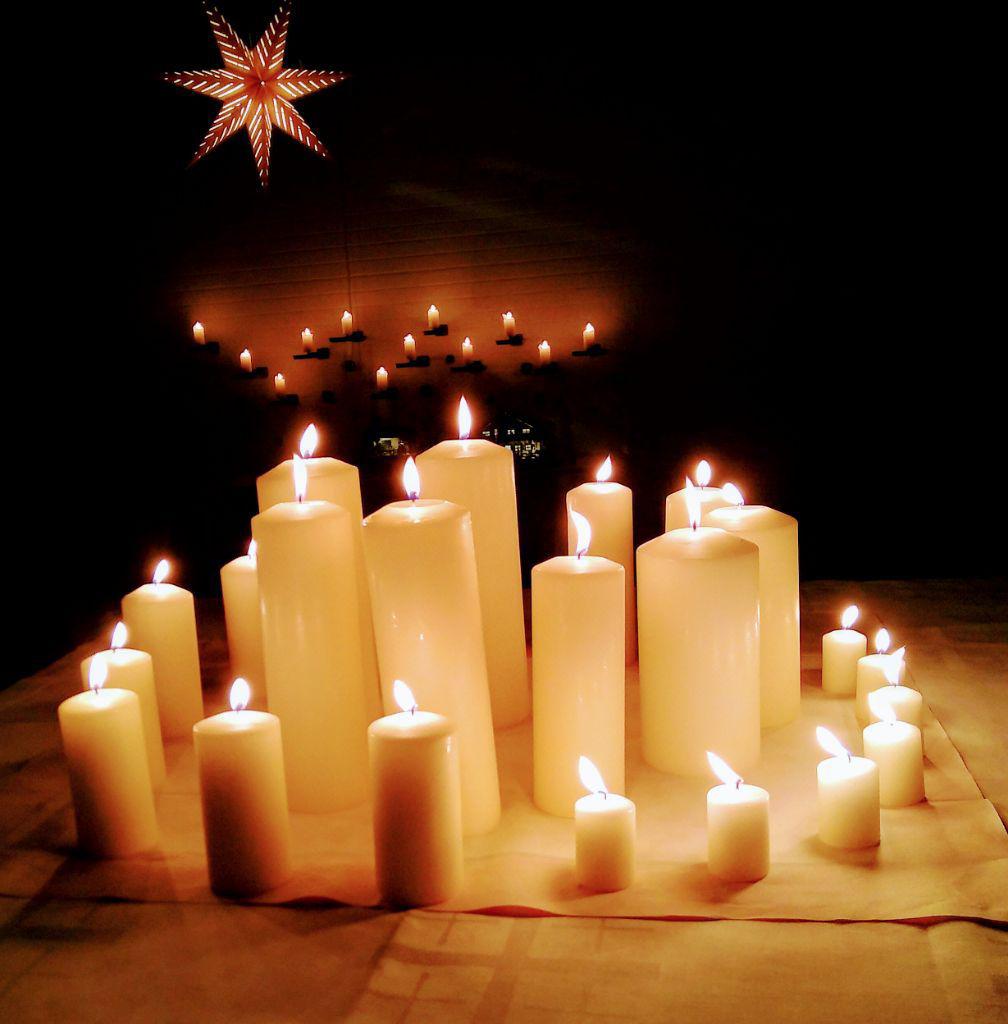 Eine Spirale aus angezündeten Kerzen im Dunkeln. Außen die kleinen, nach innen immer größer werdend. Im Hintergrund 12 Kerzen an einer Wand. Oben links ein 7-zackiger Stern.