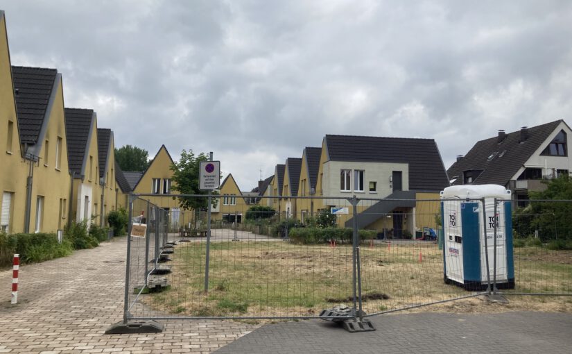 Ein eingezäuntes Stück Land, ein Dixie-Clo. Links eine Reihe spitzgibelige Häuser in gelb. Hinten rechts und und hinten frontal ebenfalls. Wolkiger Himmel. Vor dem Zaun gepflasterter Boden.