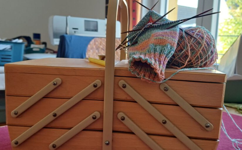 Nähkorb aus Holz, zugeklappt, oben liegt ein butes Wollknäuel mit Stricknadeln und einem Strickteil. Im Hintergrund eine Nähmaschine. Ein Fenster mit buntem Vorgang an der linken Seite.