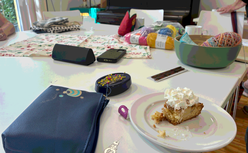 Tisch mit blauer Stofftasche links vorne, rechts vorne ein Stück Apfelkuchen mit Sahne. Auf dem Tisch verteilt_ Nähutensilien.